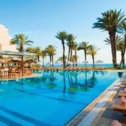Großer Außenpool mit zahlreichen Liegen, Palmen und einer stilvollen Poolbar direkt am Wasser.