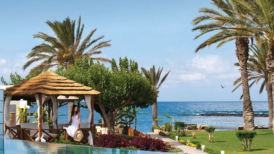 Gepflegter Garten mit Palmen und Pavillon direkt am Meer, im Hintergrund das ruhige Wasser unter blauem Himmel.