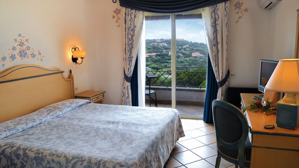 Helles Hotelzimmer mit Doppelbett, Schreibtisch und Balkon – liebevolle florale Details an Wänden und Vorhängen, Blick auf grüne Hügellandschaft.