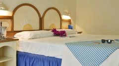 Gemütliches Doppelzimmer mit mediterranem Flair – dekoratives Holzbett mit Sonnenmotiv, frischen Blumen auf dem Bett und Urlaubsaccessoires wie Sonnenbrille und Reiseführer.