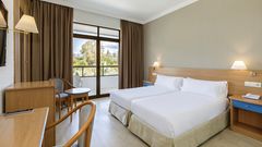 Helles Hotelzimmer mit zwei Einzelbetten, Schreibtisch, Sitzbereich und Zugang zu einem Balkon mit Blick auf eine grüne Gartenlandschaft.
