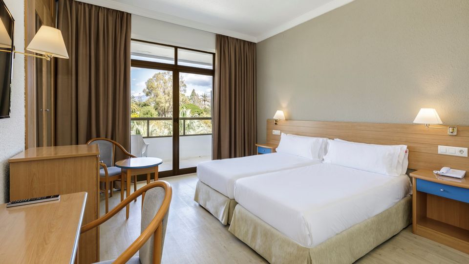 Helles Hotelzimmer mit zwei Einzelbetten, Schreibtisch, Sitzbereich und Zugang zu einem Balkon mit Blick auf eine grüne Gartenlandschaft.