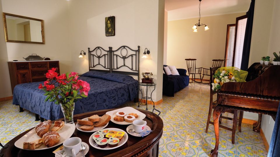 Gemütliches Doppelzimmer mit traditionellem Mobiliar, gemustertem Fliesenboden und separatem Sitzbereich – auf dem Tisch ein liebevoll angerichtetes Frühstückstablett mit Kaffee, Gebäck und frischen Blumen.