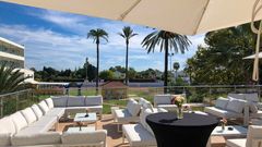 Moderne Lounge-Terrasse mit weißen Sitzgruppen und Sonnenschirmen, Blick auf Palmen und Gartenanlage unter blauem Himmel.