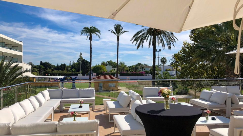 Moderne Lounge-Terrasse mit weißen Sitzgruppen und Sonnenschirmen, Blick auf Palmen und Gartenanlage unter blauem Himmel.