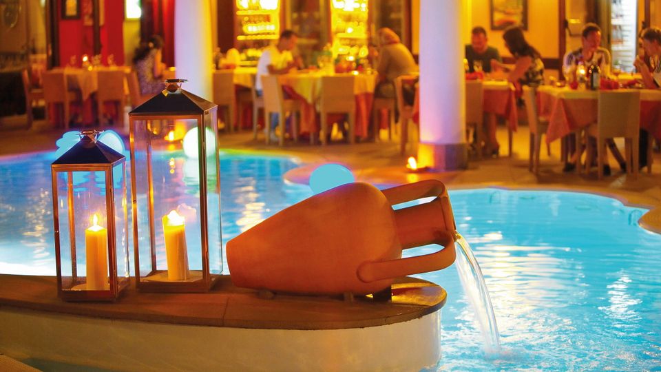 Stimmungsvoll beleuchteter Poolbereich mit Wasserspiel in Tonkrugform und dekorativen Laternen, im Hintergrund ein elegantes Restaurant mit Gästen bei Abendessen.