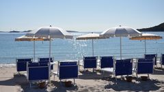 Reihe stilvoller blauer Liegestühle mit weißen Sonnenschirmen am ruhigen Sandstrand – mit Blick auf das glitzernde Meer und ferne Inseln unter klarem Himmel.