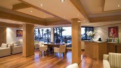Moderne Lounge mit gemütlichen Sesseln, Holzdekor und Barbereich, großer Fensterfront mit Blick auf Palmen und das Meer.