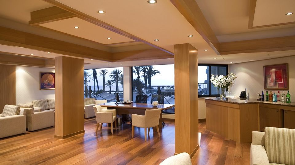 Moderne Lounge mit gemütlichen Sesseln, Holzdekor und Barbereich, großer Fensterfront mit Blick auf Palmen und das Meer.