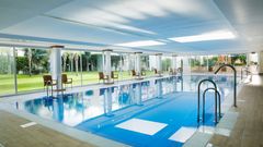 Ein moderner, überdachter Innenpool mit großen Glasfronten und Blick auf eine gepflegte Gartenanlage. Rund um den Pool stehen einzelne Stühle, die zum Verweilen einladen.