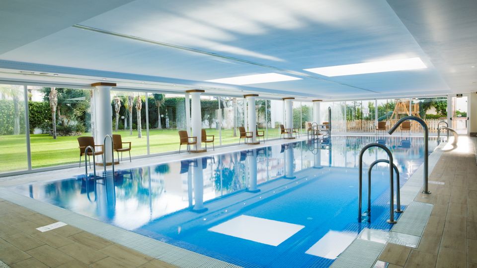 Ein moderner, überdachter Innenpool mit großen Glasfronten und Blick auf eine gepflegte Gartenanlage. Rund um den Pool stehen einzelne Stühle, die zum Verweilen einladen.