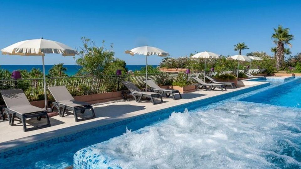 Einladender Außenpool mit Whirlpool-Bereich, Sonnenliegen und Sonnenschirmen – dahinter das glitzernde Meer und Palmen unter strahlend blauem Himmel.