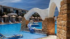 Einladender Poolbereich mit stilvollen Steinbögen, Sonnenliegen und strohgedeckten Sonnenschirmen vor zweistöckigen Hotelgebäuden – perfekte Kulisse für entspannte Urlaubstage unter strahlend blauem Himmel.