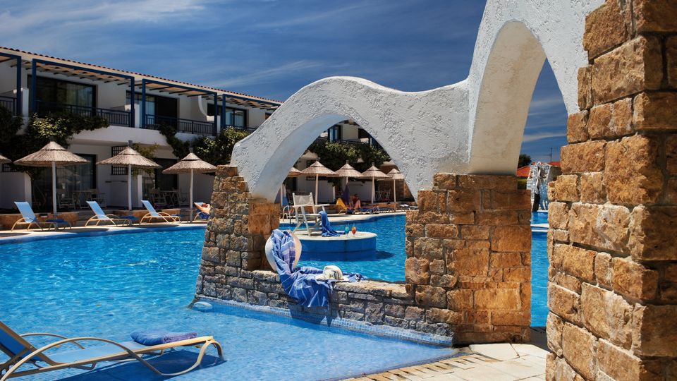 Einladender Poolbereich mit stilvollen Steinbögen, Sonnenliegen und strohgedeckten Sonnenschirmen vor zweistöckigen Hotelgebäuden – perfekte Kulisse für entspannte Urlaubstage unter strahlend blauem Himmel.