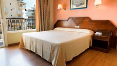 Ein einfach eingerichtetes Hotelzimmer mit einem großen Doppelbett, orangener Wand, Holzfußboden und klassischem Holzmobiliar, mit Blick aus dem Fenster auf benachbarte Gebäude.