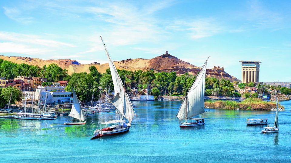 Der Nil bei Assuan: Mehrere traditionelle Segelboote (Feluccas) gleiten über das ruhige, blaue Wasser, umgeben von goldenen Wüstenhügeln und üppigem Grün entlang des Ufers.