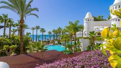 Blick über blühende Pflanzen auf eine paradiesische Hotelanlage mit weißen Türmen, tropischen Palmen, einer großzügigen Poollandschaft und dem strahlend blauen Meer im Hintergrund.