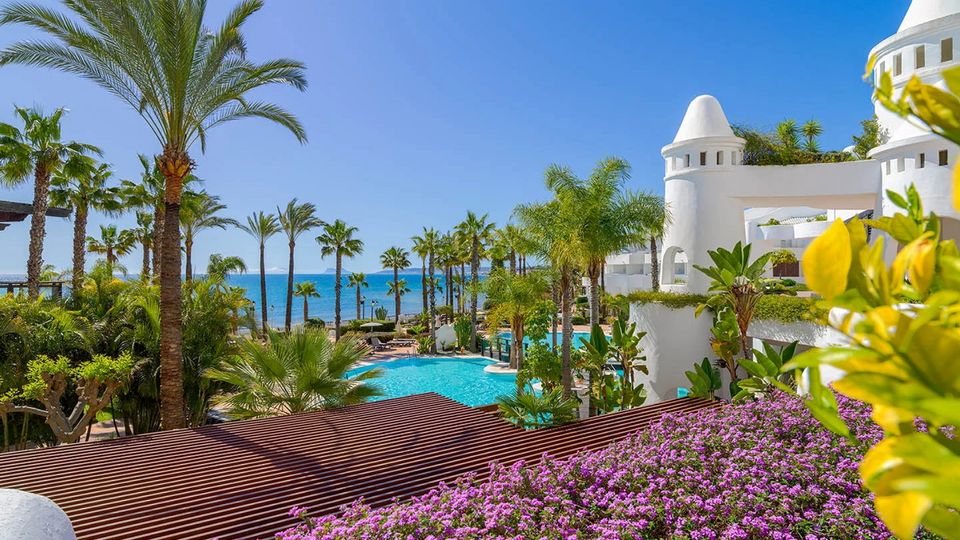Blick über blühende Pflanzen auf eine paradiesische Hotelanlage mit weißen Türmen, tropischen Palmen, einer großzügigen Poollandschaft und dem strahlend blauen Meer im Hintergrund.
