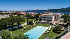 Luftaufnahme eines stilvollen Hotels mit großem Außenpool inmitten gepflegter Gartenanlagen – mit Blick auf die umliegenden Häuser, das tiefblaue Meer und die grünen Hügel Sardiniens.