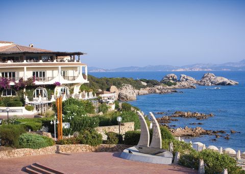Mediterranes Hotelgebäude direkt an der felsigen Küste mit traumhaftem Blick auf das türkisfarbene Meer und vorgelagerte Inseln – üppige Blumen und grüne Gartenanlagen im Vordergrund.