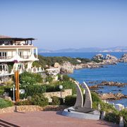 Mediterranes Hotelgebäude direkt an der felsigen Küste mit traumhaftem Blick auf das türkisfarbene Meer und vorgelagerte Inseln – üppige Blumen und grüne Gartenanlagen im Vordergrund.