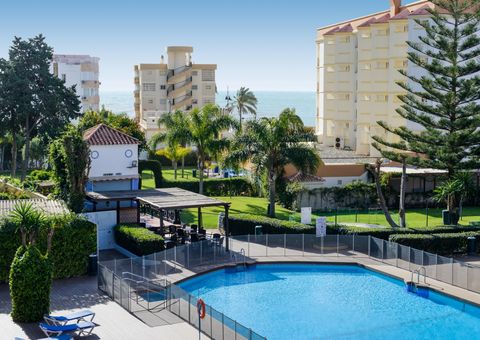 Blick auf einen runden Hotelpool mit Sicherheitszaun, daneben eine überdachte Sitzterrasse, üppige grüne Gartenanlagen mit Palmen sowie mehrstöckige Gebäude im Hintergrund, dahinter das Meer.