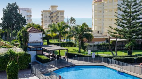 Blick auf einen runden Hotelpool mit Sicherheitszaun, daneben eine überdachte Sitzterrasse, üppige grüne Gartenanlagen mit Palmen sowie mehrstöckige Gebäude im Hintergrund, dahinter das Meer.