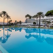 Weitläufiger Hotelpool im Abendlicht mit spiegelnden Palmen, weißen Liegen und Blick auf ein elegantes Resortgebäude vor untergehender Sonne.