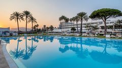 Weitläufiger Hotelpool im Abendlicht mit spiegelnden Palmen, weißen Liegen und Blick auf ein elegantes Resortgebäude vor untergehender Sonne.