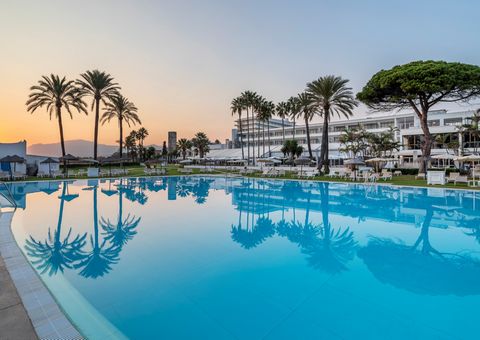 Weitläufiger Hotelpool im Abendlicht mit spiegelnden Palmen, weißen Liegen und Blick auf ein elegantes Resortgebäude vor untergehender Sonne.