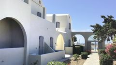 Ein gepflegter Gartenweg führt durch die hellen Gebäude im typisch kykladischen Stil mit weißen Fassaden und grauen Fensterläden – im Hintergrund lädt der Poolbereich zum Entspannen ein.