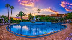 Großer Hotelpool mit Palmenkulisse bei Sonnenuntergang – das Wasser spiegelt den farbenprächtigen Himmel, umrahmt von Liegestühlen und mediterranem Ambiente.