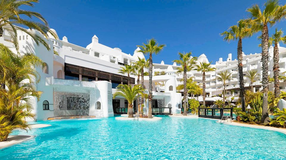 Weitläufige Poollandschaft eines weißen Luxusresorts mit Palmen, Wasserfällen und türkisblauem Wasser – perfekte Kulisse für einen entspannten Urlaub in der Sonne.