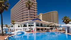 Modernes Hotelgebäude mit mehreren Etagen und geschwungenen Balkonen, direkt an einem großen Außenpool mit Liegestühlen und Sonnenschirmen, unter strahlend blauem Himmel – ideal für einen sonnigen Badeurlaub.