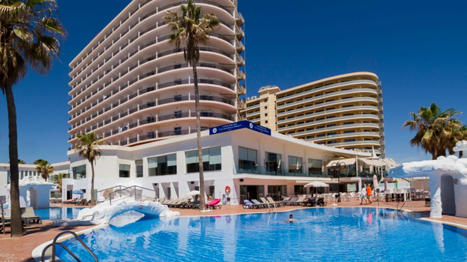 Modernes Hotelgebäude mit mehreren Etagen und geschwungenen Balkonen, direkt an einem großen Außenpool mit Liegestühlen und Sonnenschirmen, unter strahlend blauem Himmel – ideal für einen sonnigen Badeurlaub.