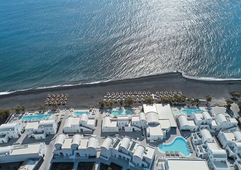 Luftaufnahme des Costa Grand Resort & Spa auf Santorin – elegante Hotelanlage direkt am schwarzen Lavastrand von Kamari mit mehreren Pools, weißen Gebäuden im Kykladenstil und herrlichem Blick auf das funkelnde Meer.