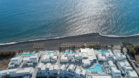 Luftaufnahme des Costa Grand Resort & Spa auf Santorin – elegante Hotelanlage direkt am schwarzen Lavastrand von Kamari mit mehreren Pools, weißen Gebäuden im Kykladenstil und herrlichem Blick auf das funkelnde Meer.
