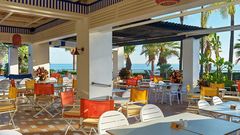 Heller, farbenfroher Außenbereich eines Restaurants mit Blick auf das Meer, umgeben von Palmen und tropischer Vegetation.