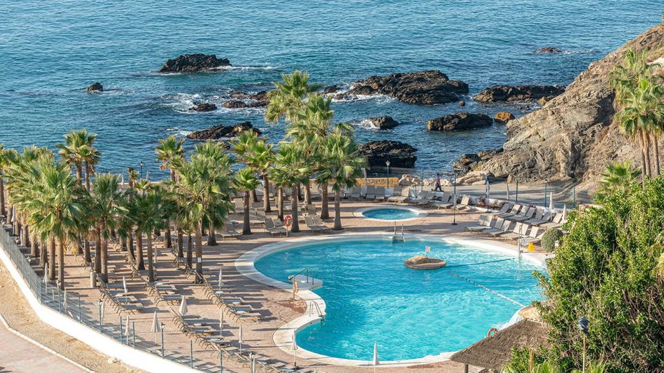 Großzügige Poolanlage direkt an der felsigen Küste mit zahlreichen Palmen, Liegestühlen und einem separaten Whirlpool – perfekter Ort zum Entspannen mit Blick aufs Meer.
