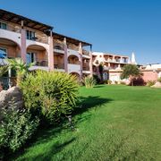 Hotelanlage im mediterranen Stil mit rosa-ockerfarbenen Fassaden, Balkonen und gepflegter Gartenanlage – umgeben von Palmen, Büschen und großen Felsen unter strahlend blauem Himmel.