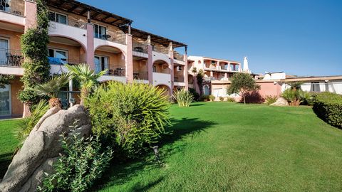 Hotelanlage im mediterranen Stil mit rosa-ockerfarbenen Fassaden, Balkonen und gepflegter Gartenanlage – umgeben von Palmen, Büschen und großen Felsen unter strahlend blauem Himmel.