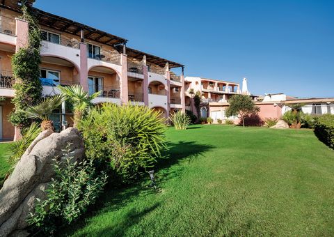 Hotelanlage im mediterranen Stil mit rosa-ockerfarbenen Fassaden, Balkonen und gepflegter Gartenanlage – umgeben von Palmen, Büschen und großen Felsen unter strahlend blauem Himmel.