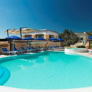 Einladender Außenpool mit türkisblauem Wasser, umgeben von blauen Sonnenschirmen und Liegestühlen vor einer mediterranen Hotelanlage unter klarem Himmel.