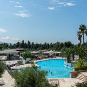 Moderner Hotelpoolbereich mit großer, geschwungener Poolanlage, umgeben von Sonnenliegen, Palmen und mediterraner Vegetation – im Hintergrund flache Gebäude, Pinien und blauer Himmel.