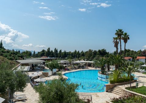 Moderner Hotelpoolbereich mit großer, geschwungener Poolanlage, umgeben von Sonnenliegen, Palmen und mediterraner Vegetation – im Hintergrund flache Gebäude, Pinien und blauer Himmel.