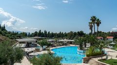 Moderner Hotelpoolbereich mit großer, geschwungener Poolanlage, umgeben von Sonnenliegen, Palmen und mediterraner Vegetation – im Hintergrund flache Gebäude, Pinien und blauer Himmel.