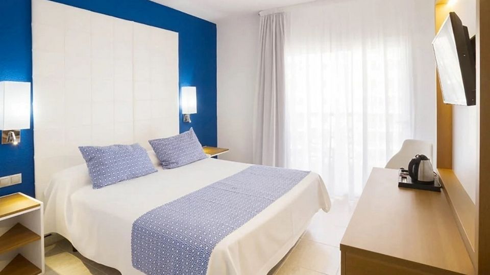 Helles Doppelzimmer mit blau-weißem Farbschema, großem Bett mit zwei blauen Zierkissen, weißen Vorhängen vor dem Balkon und einem Schreibtisch mit Wasserkocher und Flachbildfernseher.