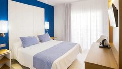 Helles Doppelzimmer mit blau-weißem Farbschema, großem Bett mit zwei blauen Zierkissen, weißen Vorhängen vor dem Balkon und einem Schreibtisch mit Wasserkocher und Flachbildfernseher.