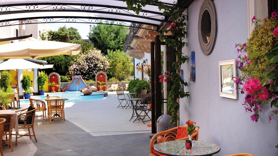Stilvoll gestalteter Innenhof mit kleinem Brunnen, bunten Wanddekorationen, gemütlichen Sitzplätzen und mediterraner Bepflanzung – lebendige Farben und liebevolle Details sorgen für ein einladendes Ambiente.