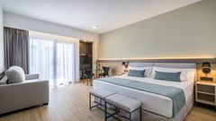 Modernes, geräumiges Hotelzimmer mit Kingsize-Bett, hellen Holzböden, gemütlicher Sitzecke, Schreibtisch und bodentiefen Fenstern, die viel Tageslicht hereinlassen.